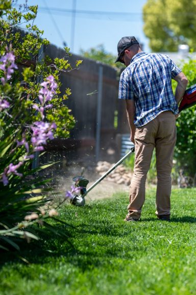 Mann nutzt eine Motorsense im Garten, um Gras zu trimmen, umgeben von Blumen.