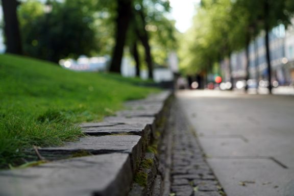 Grünstreifen neben einem gepflasterten Weg, umgeben von Bäumen und Gebäuden.