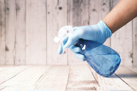 Hand mit blauer Gummihandschuh hält eine Sprühflasche auf einem Holzuntergrund.