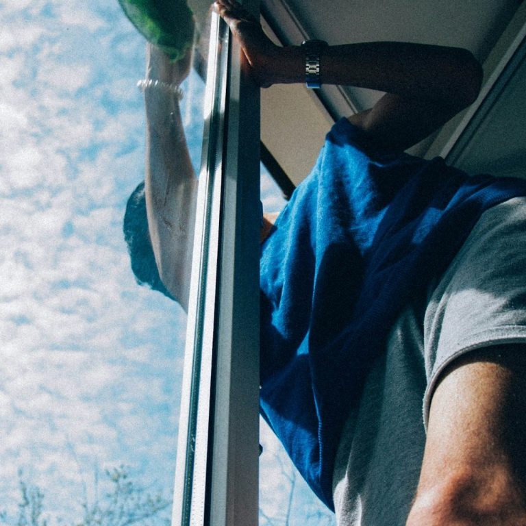 Eine Person putzt ein Fenster von innen, während sie auf einen Stuhl steht.
